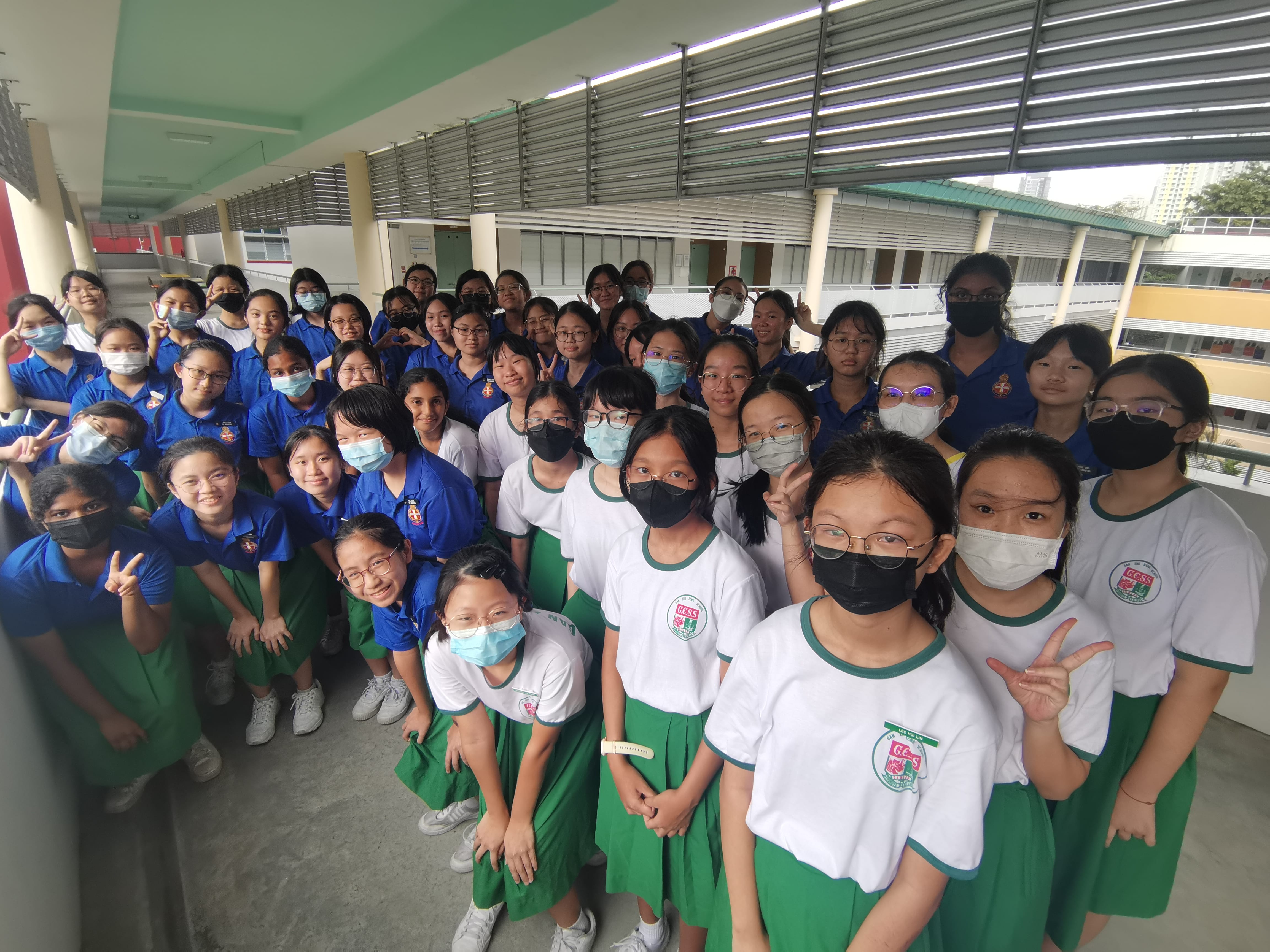 Large group of students in school uniforms in a corridor, many wearing masks and making peace signs.